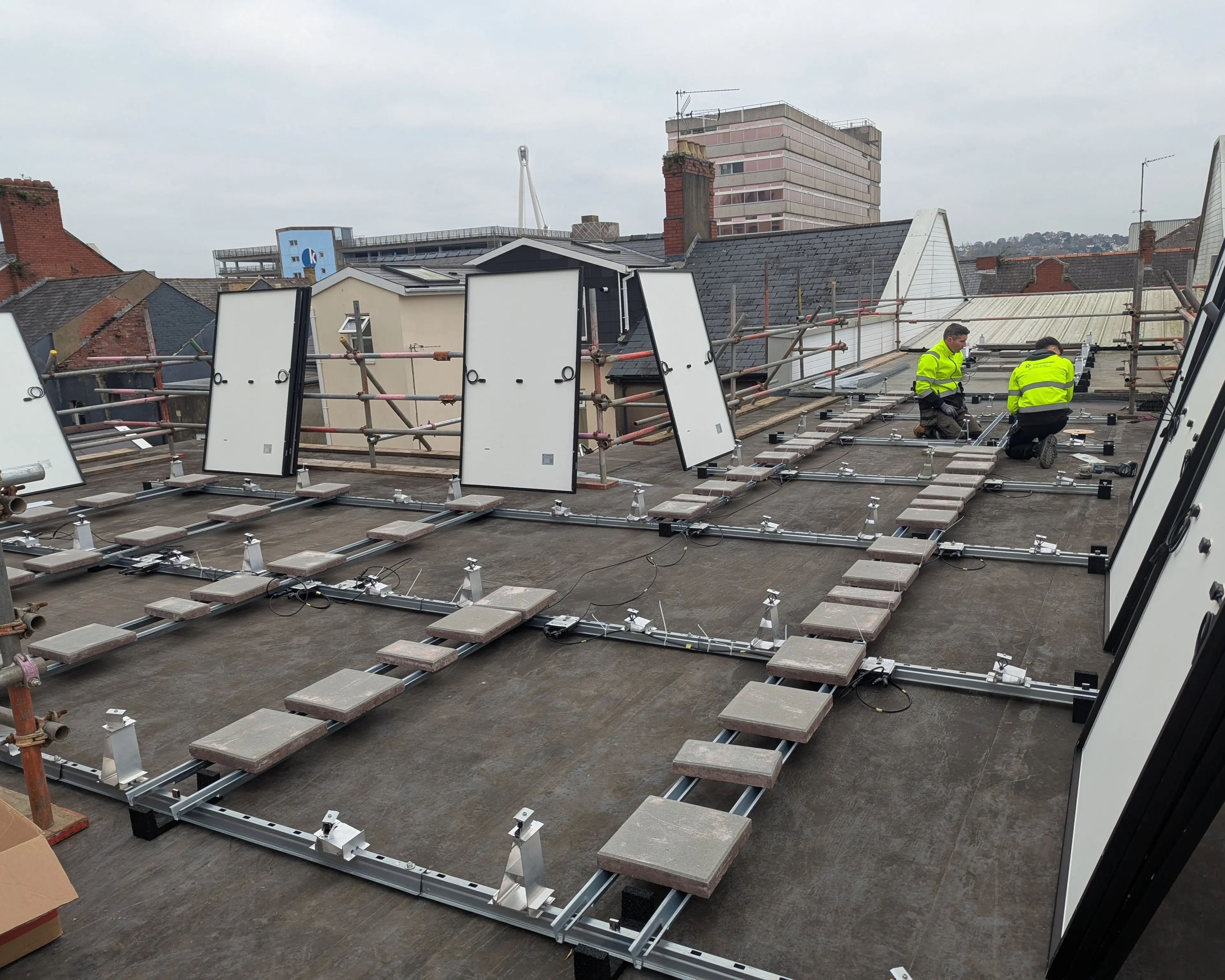 Two installers fitting the ballasted mounting frame and panels on the flat roof of iTec Skills' training centre in Newport, with the town and Newport Civic Centre in the background