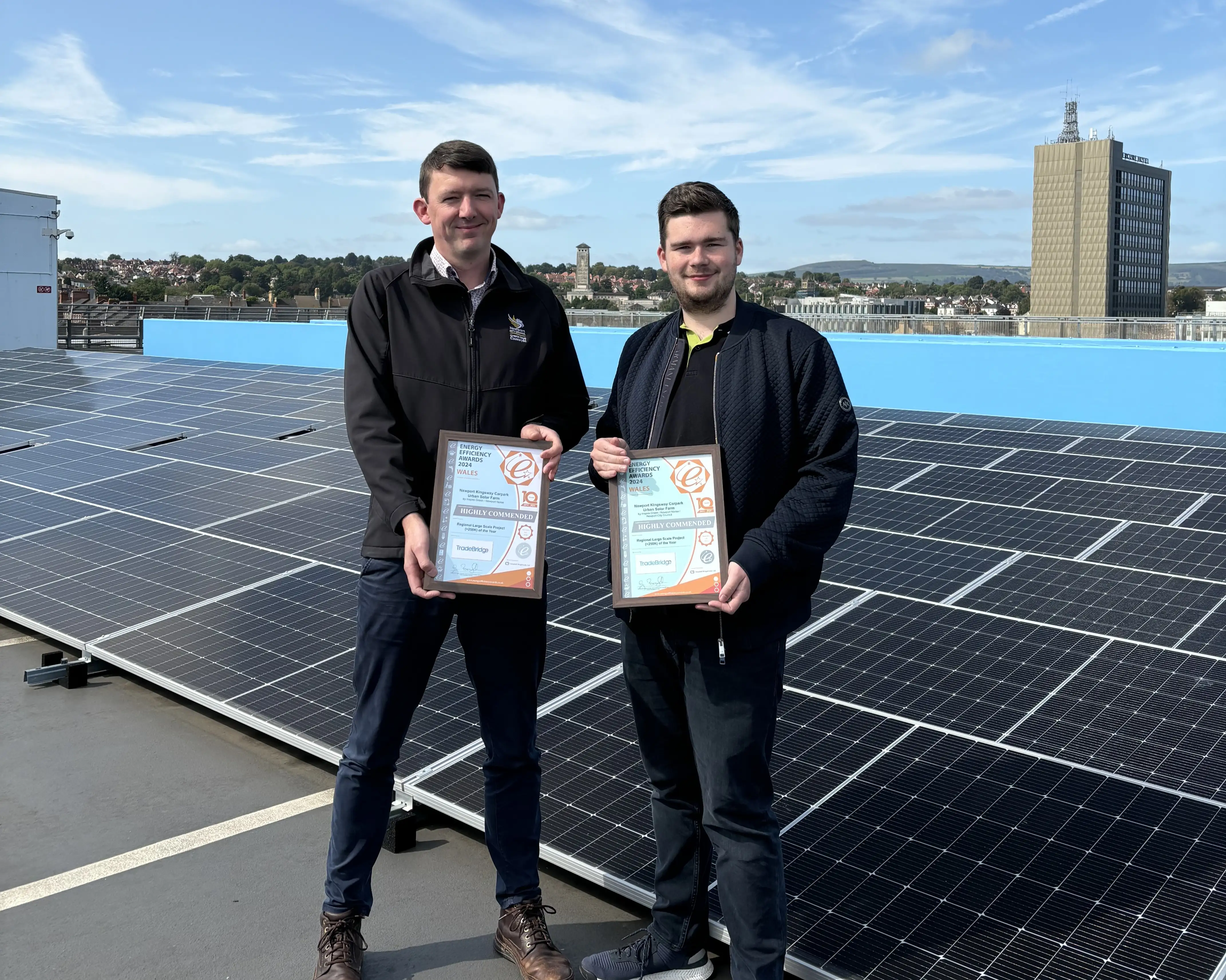 Two men holding Highly Commended certificates from the 2024 Wales Energy Efficiency Awards on the roof of Newport Kingsway car park, with the completed solar array behind them