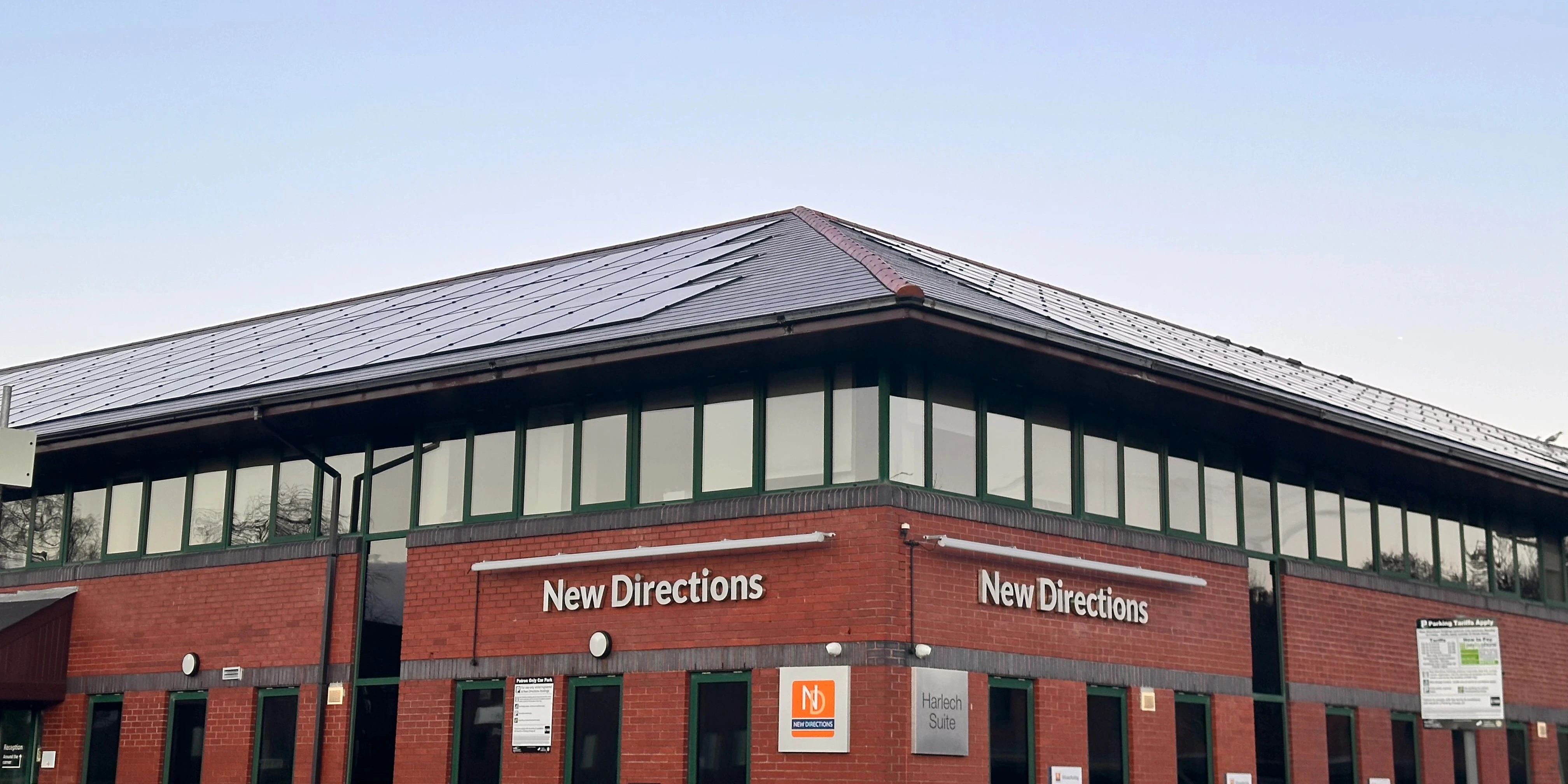 Roof-integrated solar panels across the pitched roof of the New Directions red-brick commercial building, with green-framed windows and signage below against a…Roof-integrated solar panels across the pitched roof of the New Directions red-brick commercial building, with green-framed windows and signage below against a clear sky.