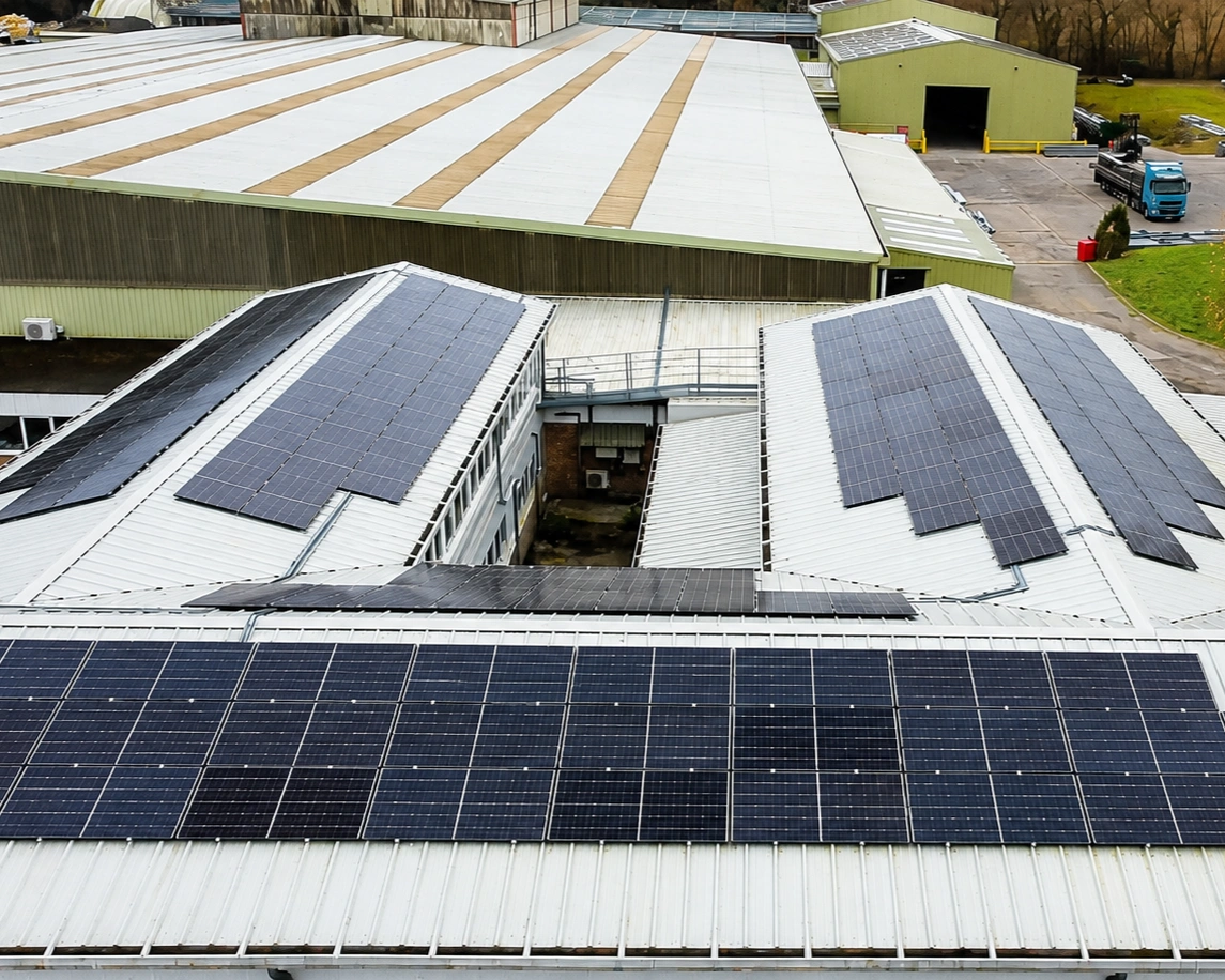 Aerial view of a commercial rooftop solar PV installation on an industrial building, with panel arrays across the pitched metal roofs and warehouses beyond.