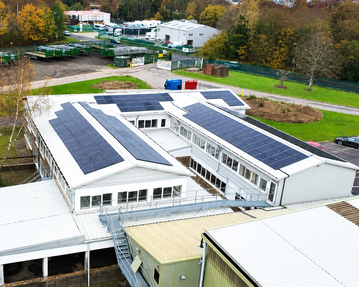 Aerial view of a commercial rooftop solar PV installation across the pitched roofs of an industrial building, with multiple panel arrays facing the yard and trailer storage.