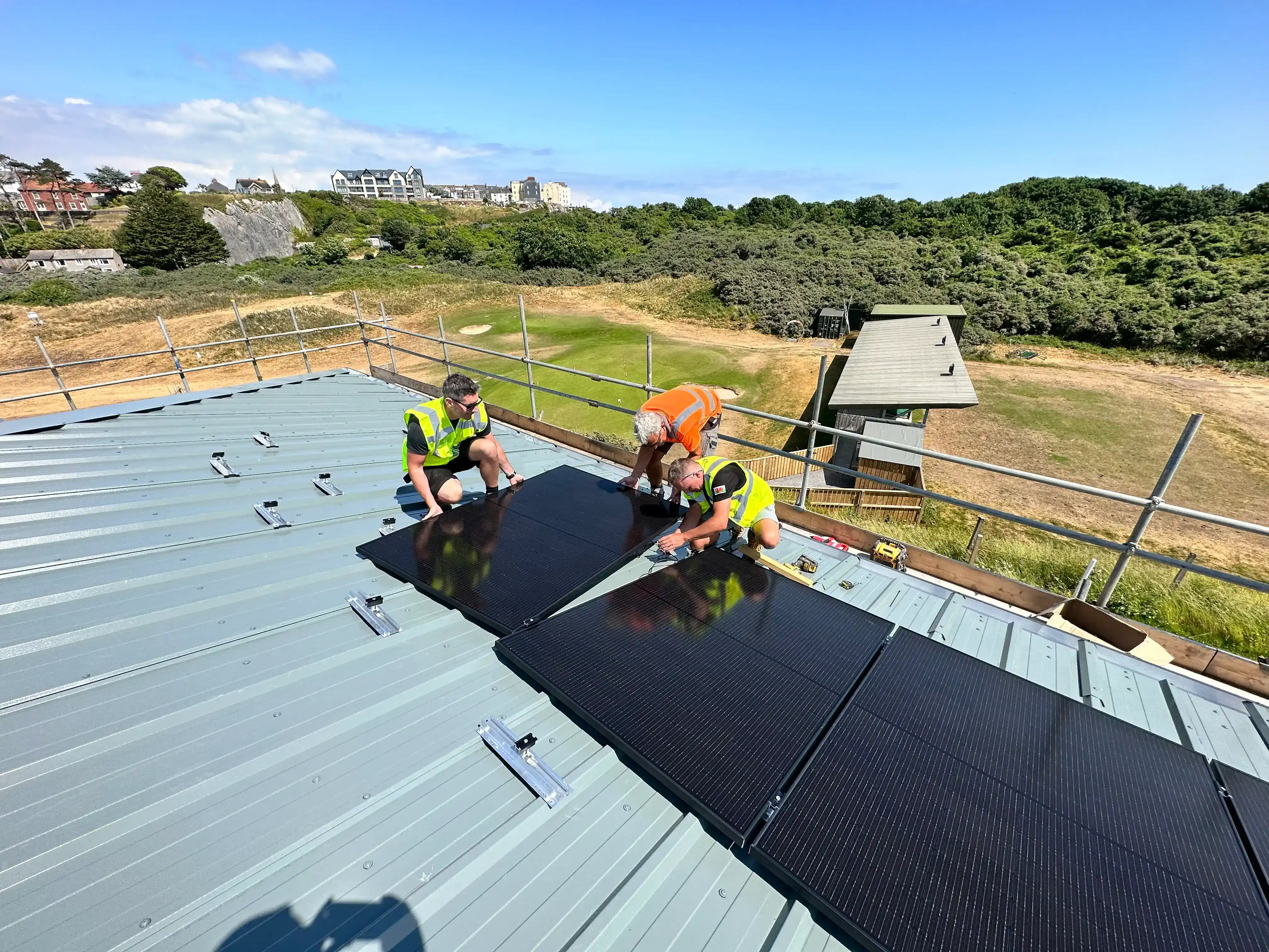 Commercial solar PV installation in progress on a golf clubhouse roof, with engineers mounting black panels on standing seam cladding.