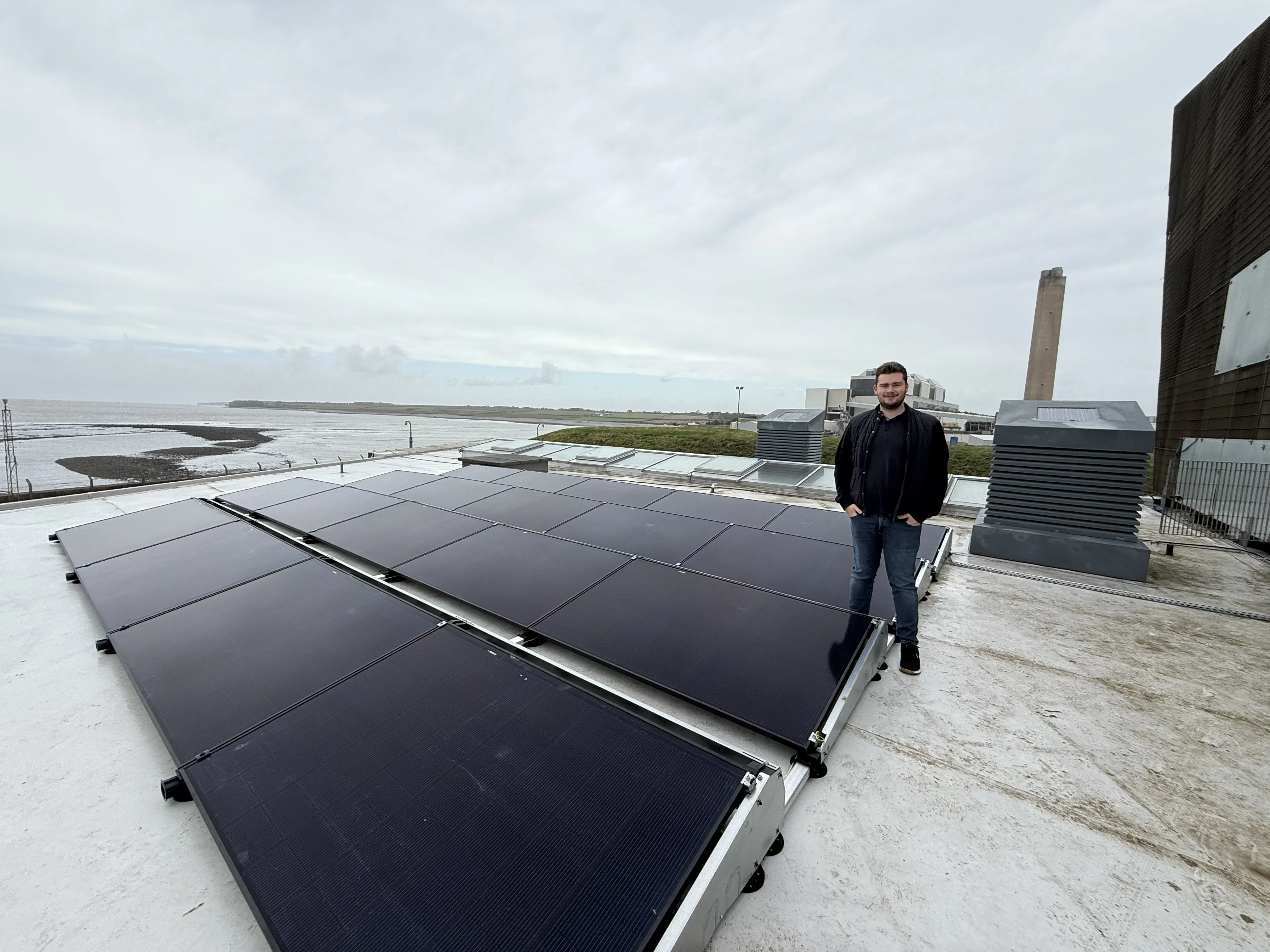 Person standing beside a flat-roof commercial solar PV array on ballasted frames at a coastal site near Aberthaw, with the estuary and a tall chimney behind.
