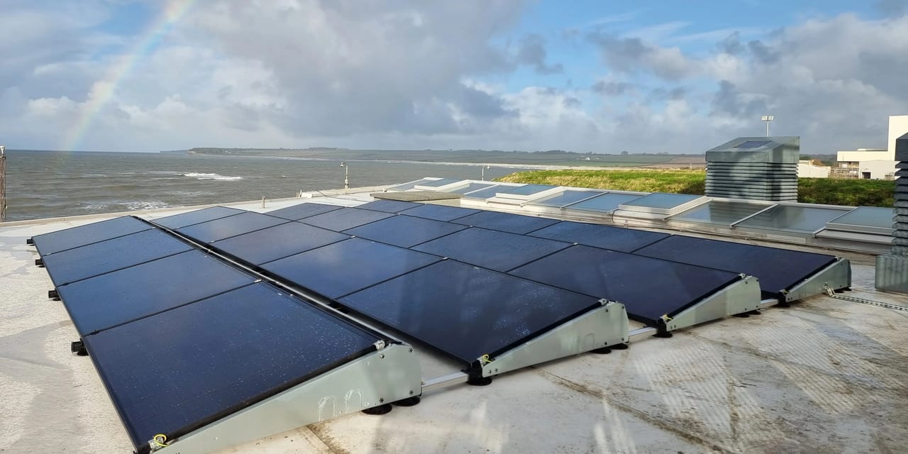 Commercial solar PV array on ballasted mounting frames across a flat coastal roof, with rooflights behind the panels and a faint rainbow over the sea.