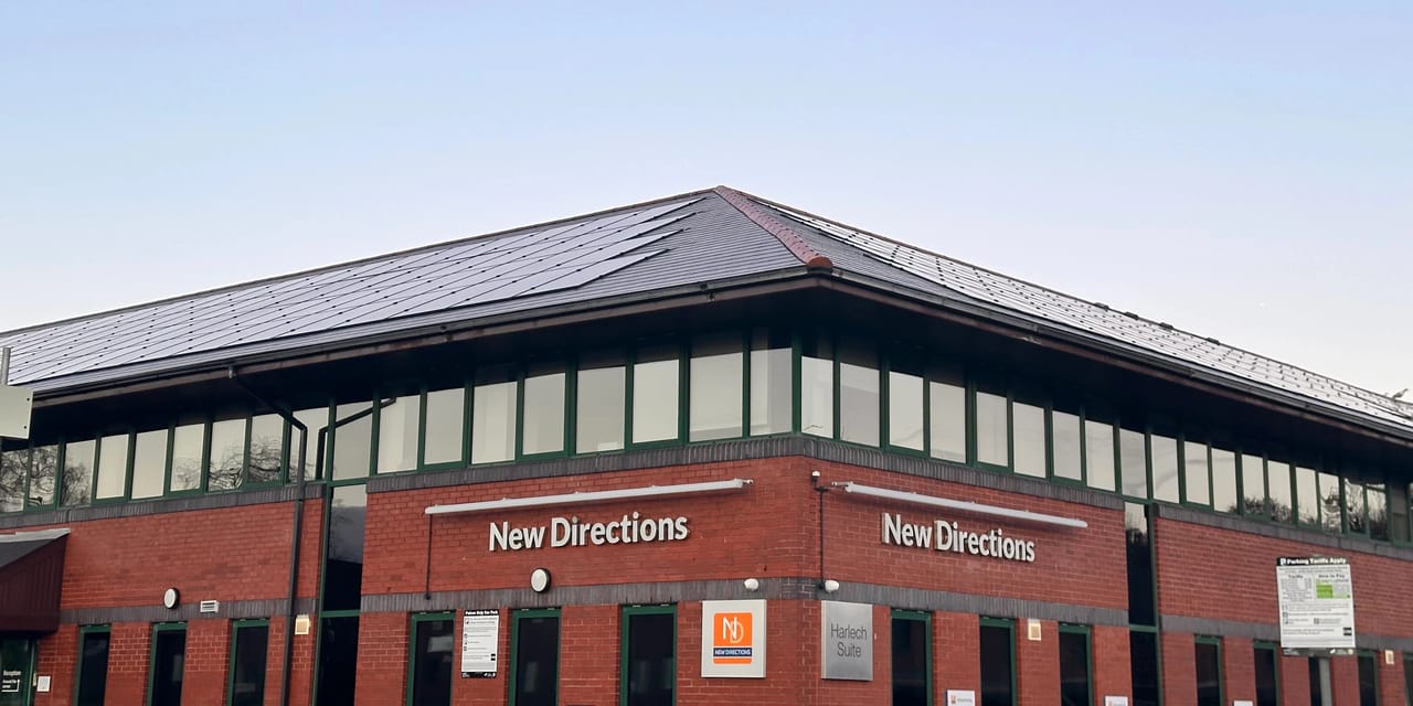 Roof-integrated solar panels across the pitched roof of the New Directions red-brick commercial building, with green-framed windows and signage below against a…Roof-integrated solar panels across the pitched roof of the New Directions red-brick commercial building, with green-framed windows and signage below against a clear sky.