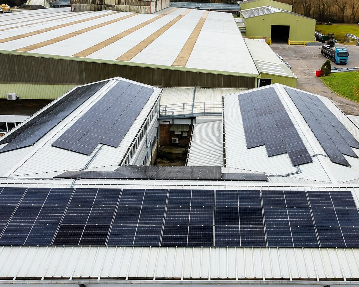 Aerial view of a commercial rooftop solar PV installation on an industrial building, with panel arrays across the pitched metal roofs and warehouses beyond.