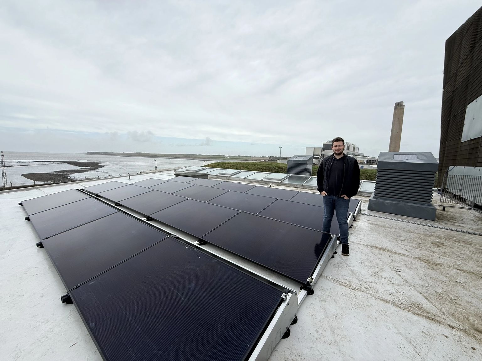 Person standing beside a flat-roof commercial solar PV array on ballasted frames at a coastal site near Aberthaw, with the estuary and a tall chimney behind.