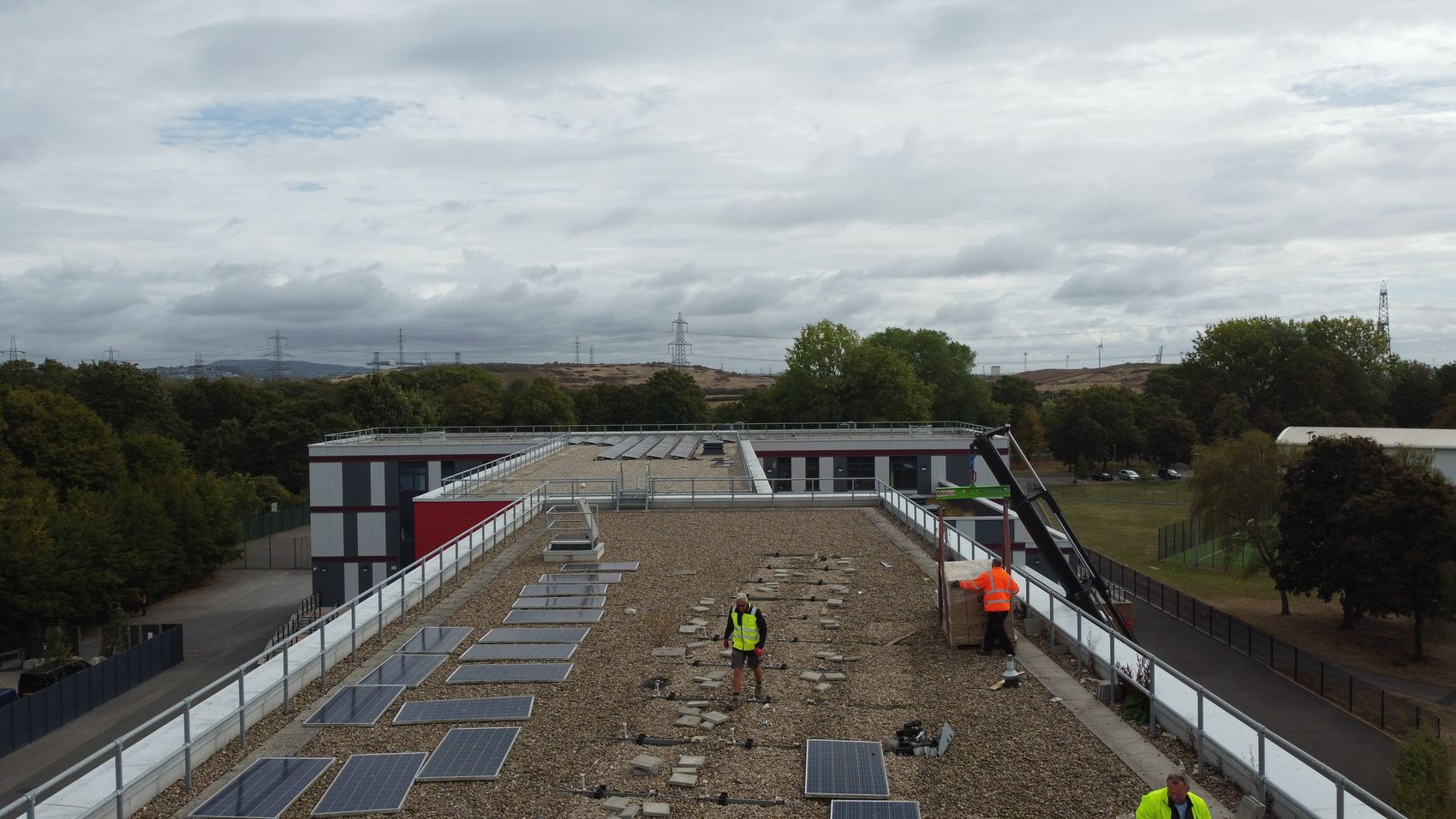 Installers fitting solar panels on the flat roof of a school building, with a crane lifting materials up during the works