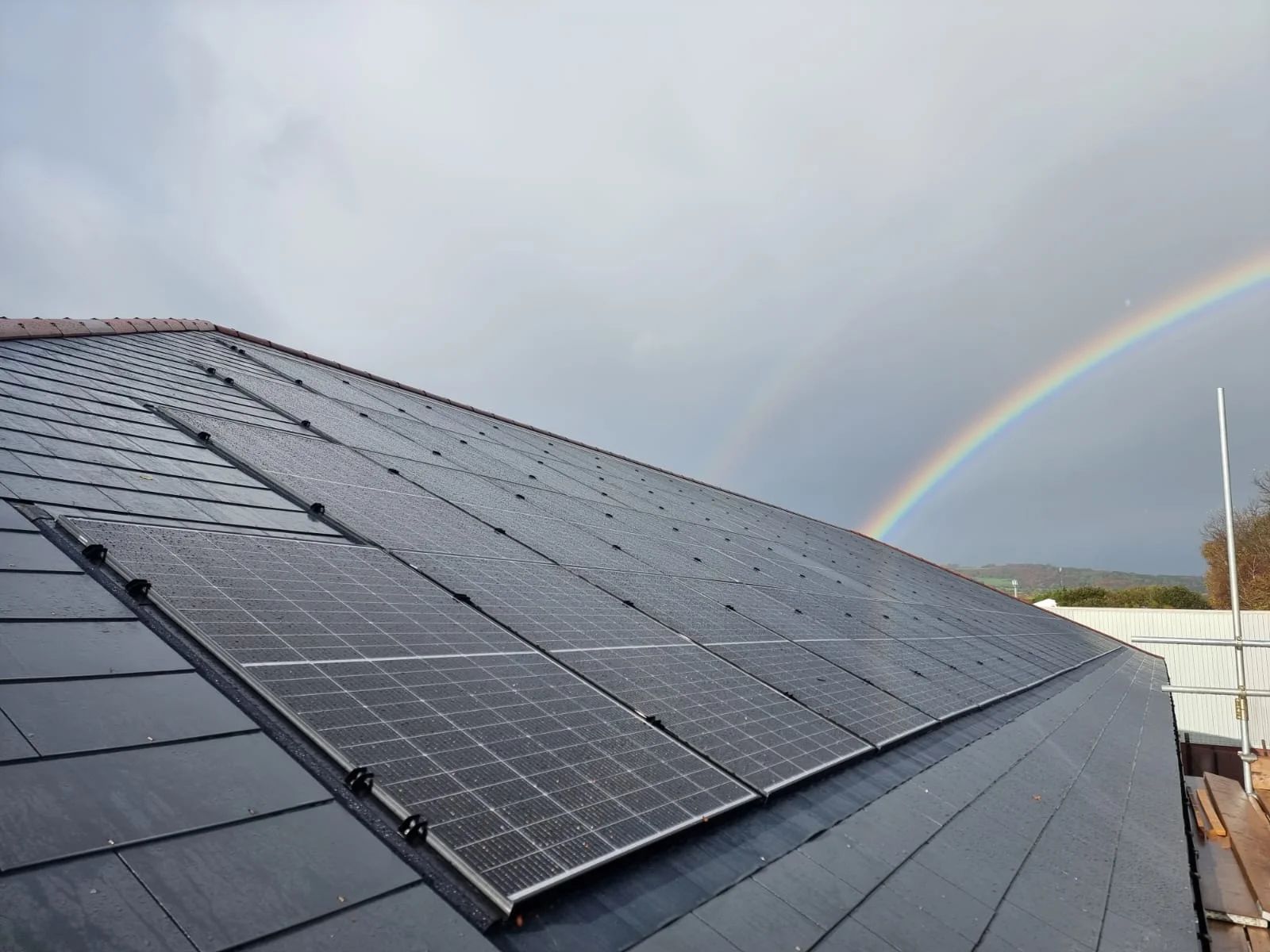 In-roof solar panels integrated flush with a dark tiled roof, with a double rainbow in the sky behind