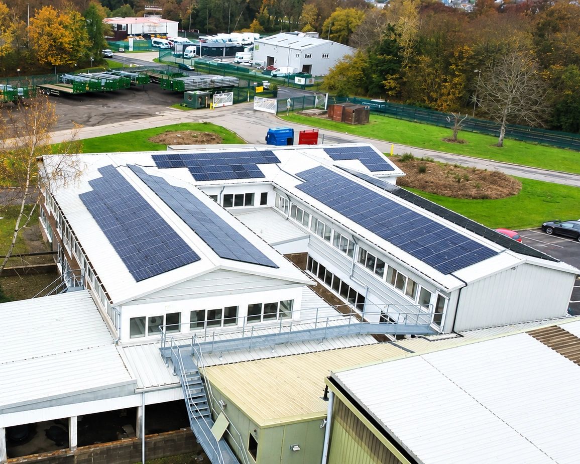 Aerial view of a commercial rooftop solar PV installation across the pitched roofs of an industrial building, with multiple panel arrays facing the yard and trailer storage.